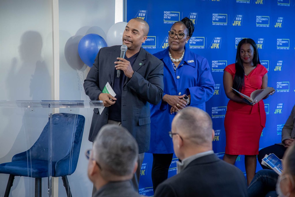 A man speaks at an event with two women attendees behind him, surrounded by decorative balloons and a blue backdrop.