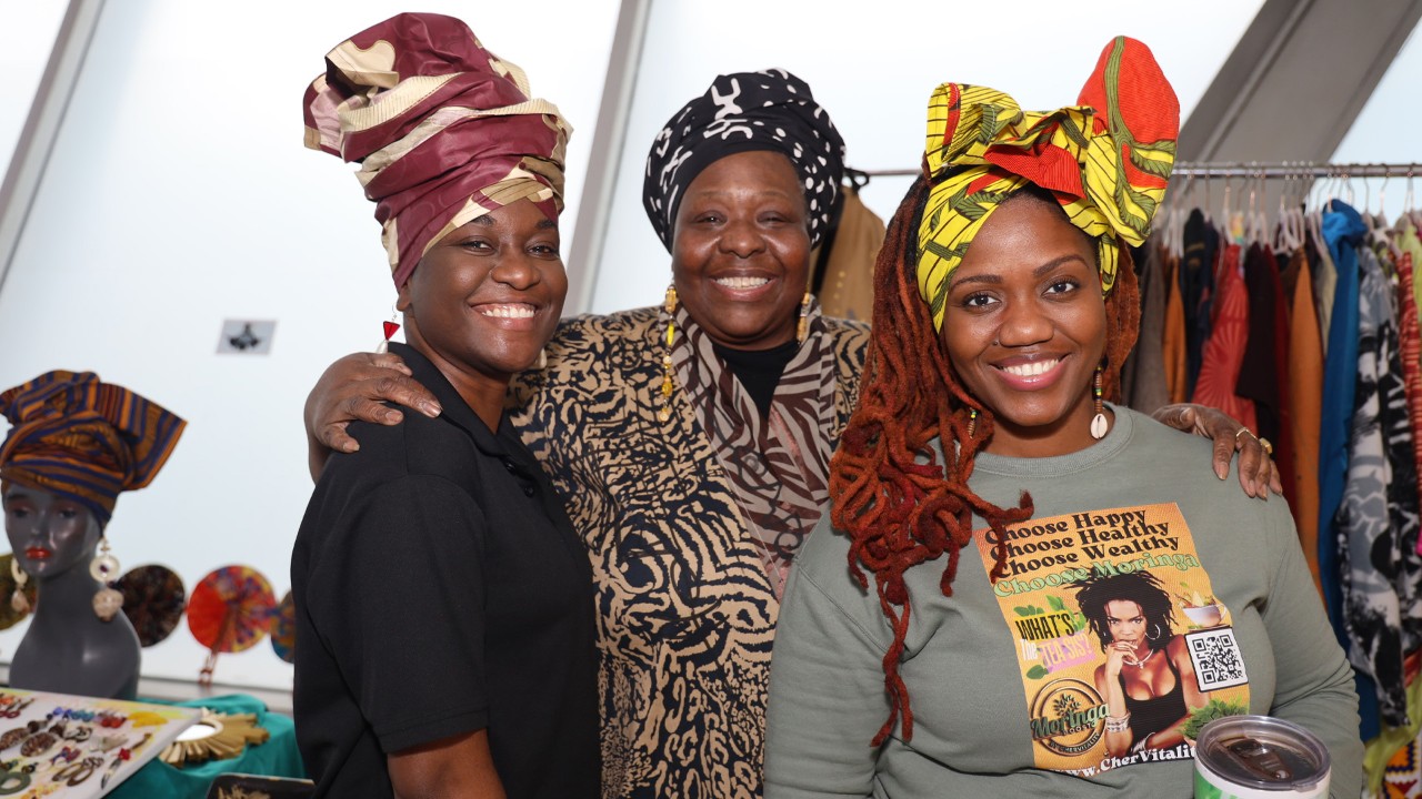 Three women in traditional headwraps smile at the camera